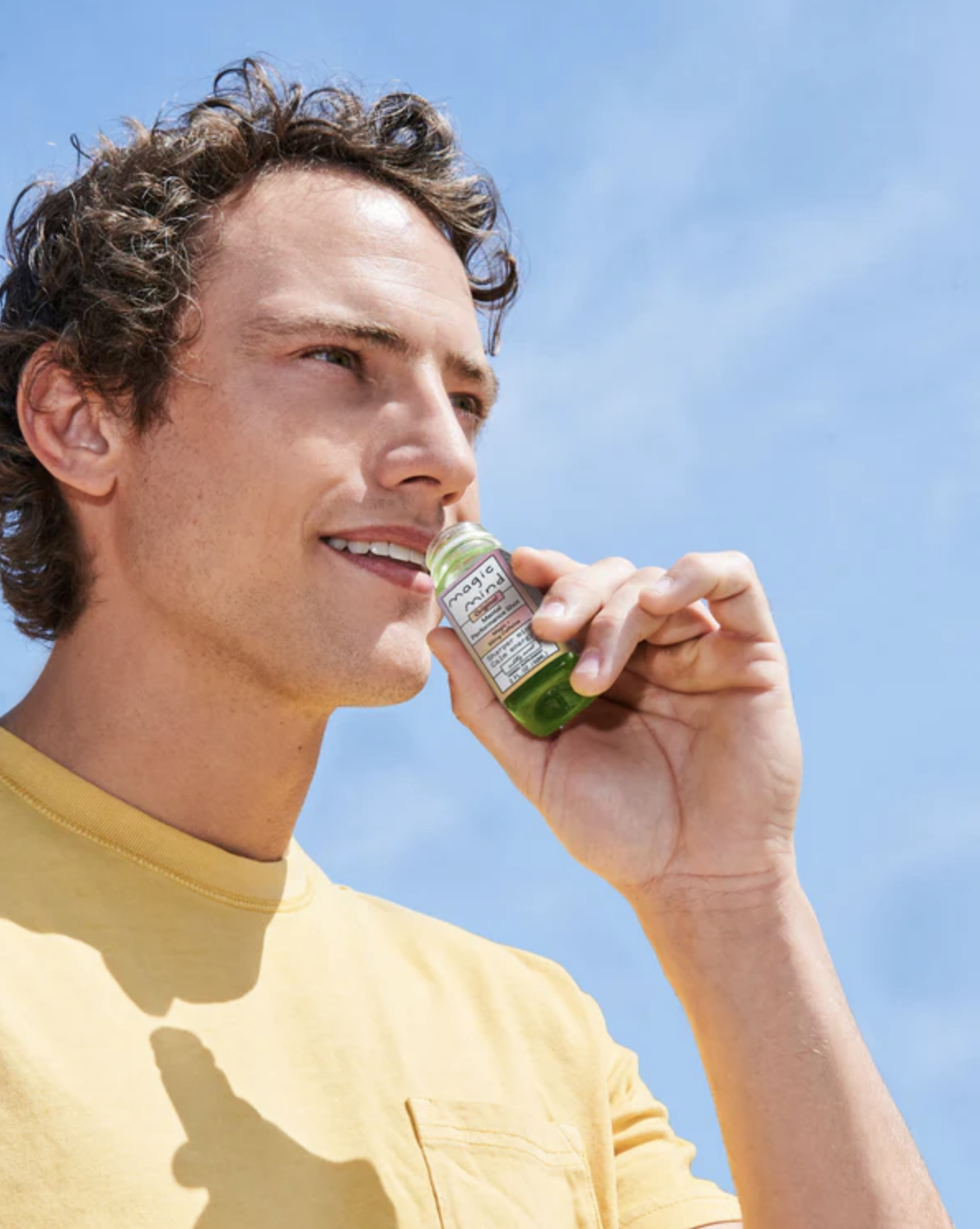 A person in a yellow shirt holds a small green bottle near their mouth against a blue sky background.