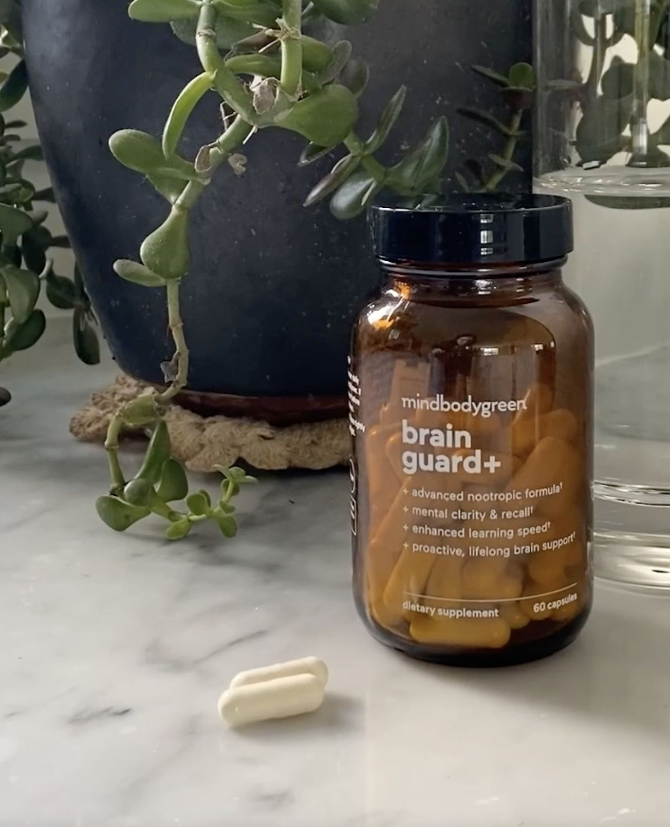 A bottle labeled "brain guard+" with yellow capsules sits on a marble surface beside two loose capsules, a glass of water, and a potted plant in the background.