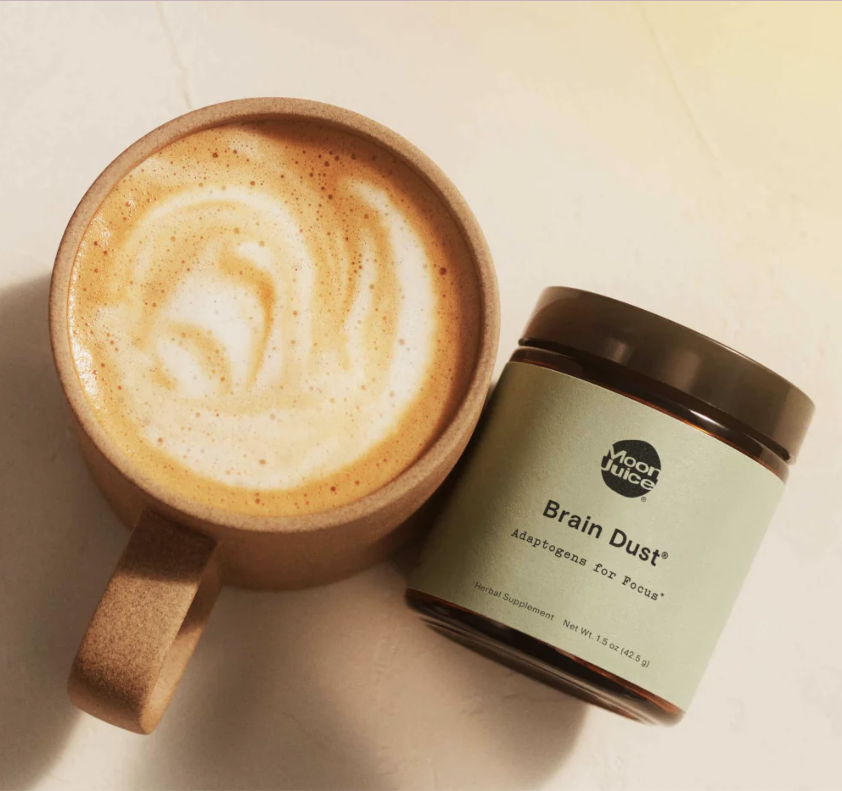 A mug of frothy coffee sits next to a jar labeled "Moon Juice Brain Dust Adaptogens for Focus" on a light-colored surface.