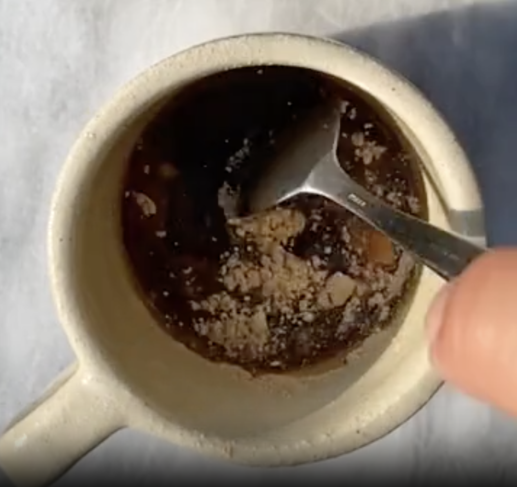 A hand holds a spoon, stirring a dark liquid with granules in a white mug placed on a light surface.