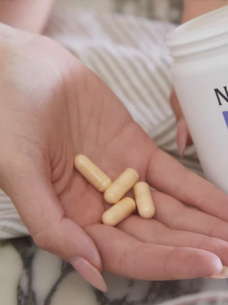 A hand holds four beige capsules next to a white plastic bottle.