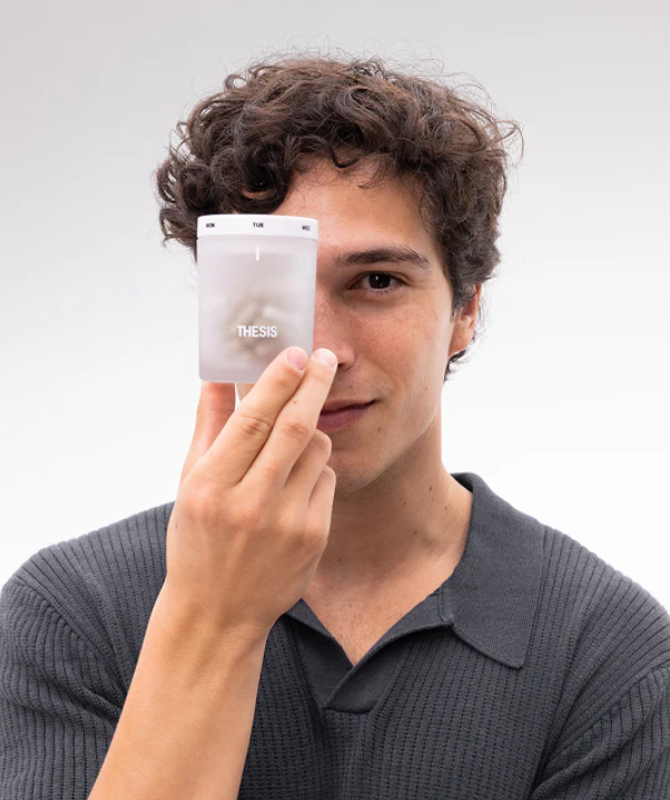 A person with curly dark hair holds a white, square container labeled "THESIS" in front of their face, partially covering it.