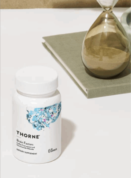 A bottle of Thorne Brain Factors dietary supplement sits on a white surface next to a sand hourglass and a closed green book.