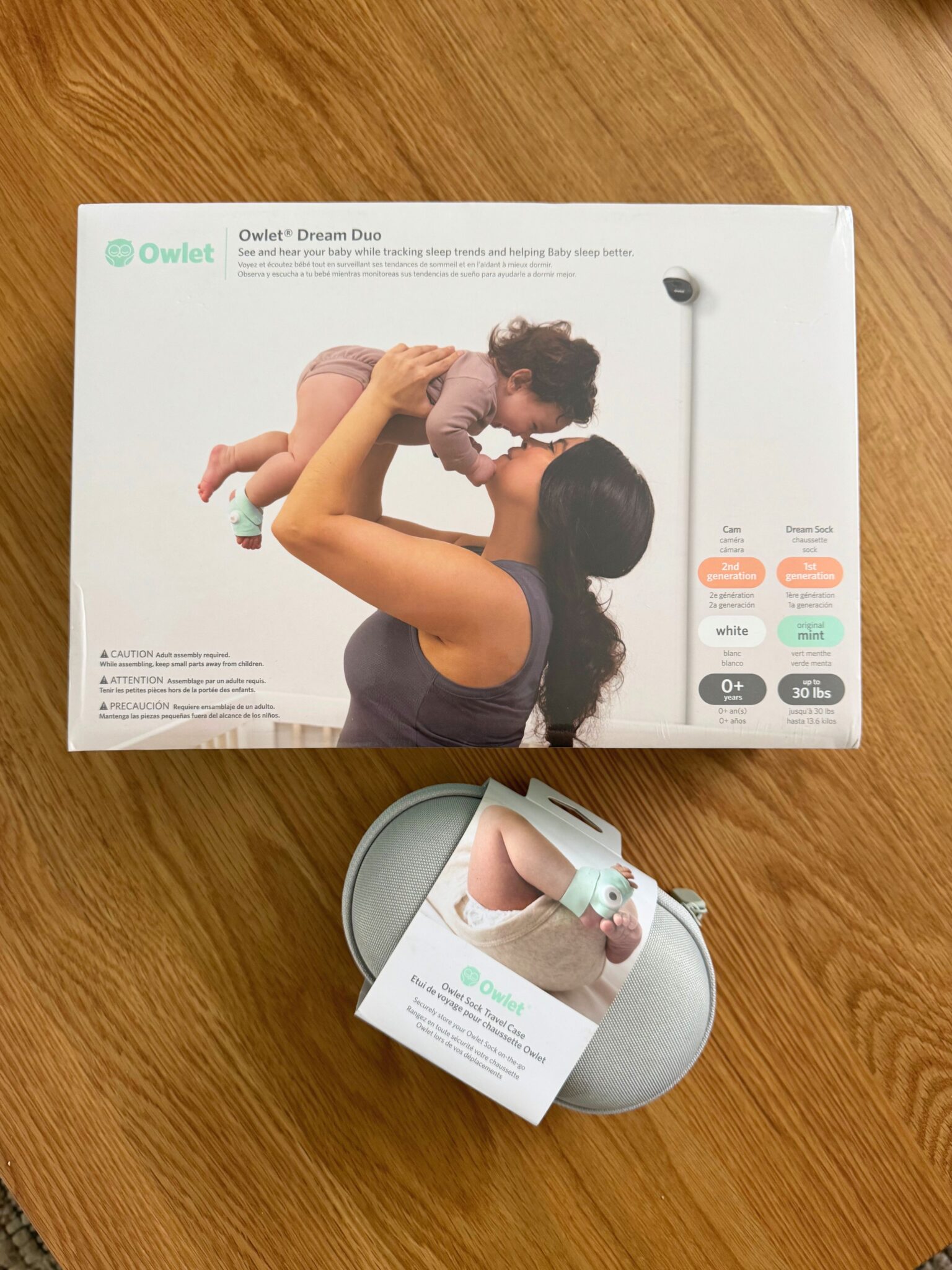 A boxed Owlet Dream Duo baby monitor and sock set sits on a wooden surface, with the product case placed in front of the box.