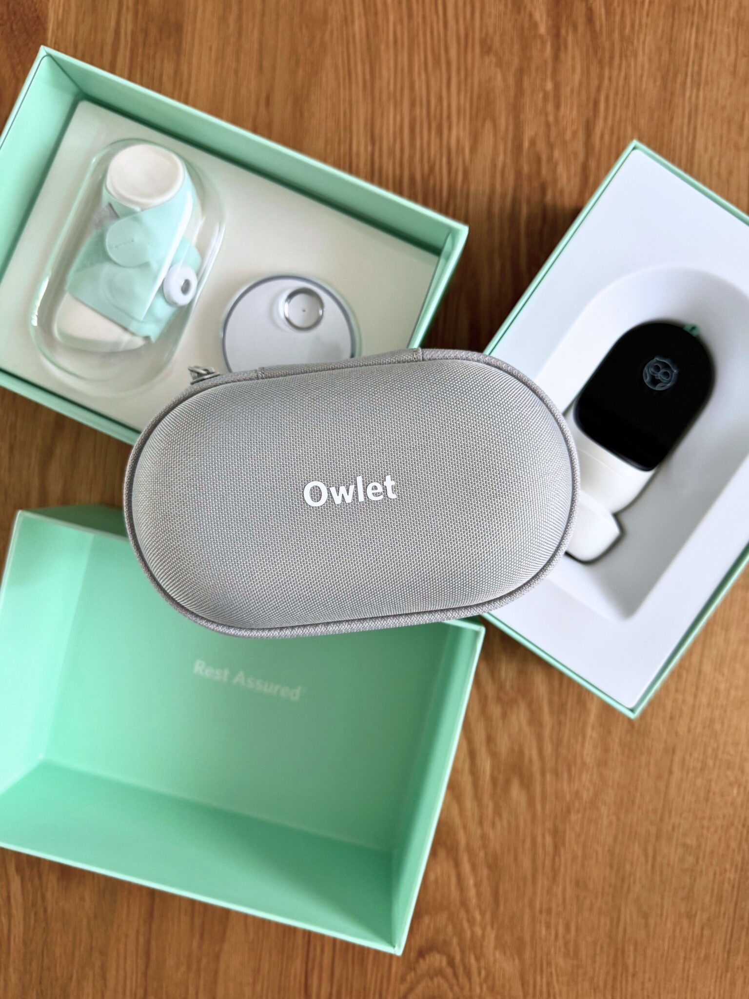 Three mint green boxes on a wooden surface contain an Owlet branded case, a sensor, and a charging base, as part of a baby monitoring device set.