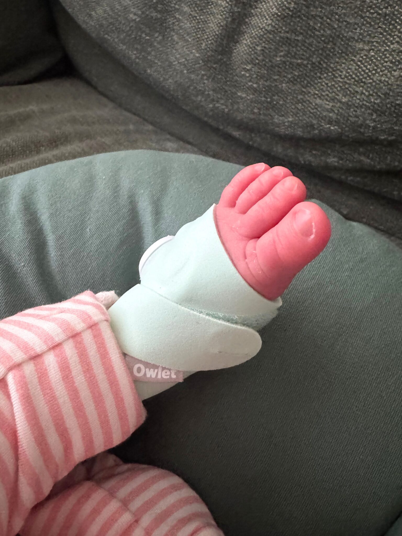A baby’s foot wearing a soft mint-green monitor rests on a cushion. The baby is dressed in pink and white striped pajamas.