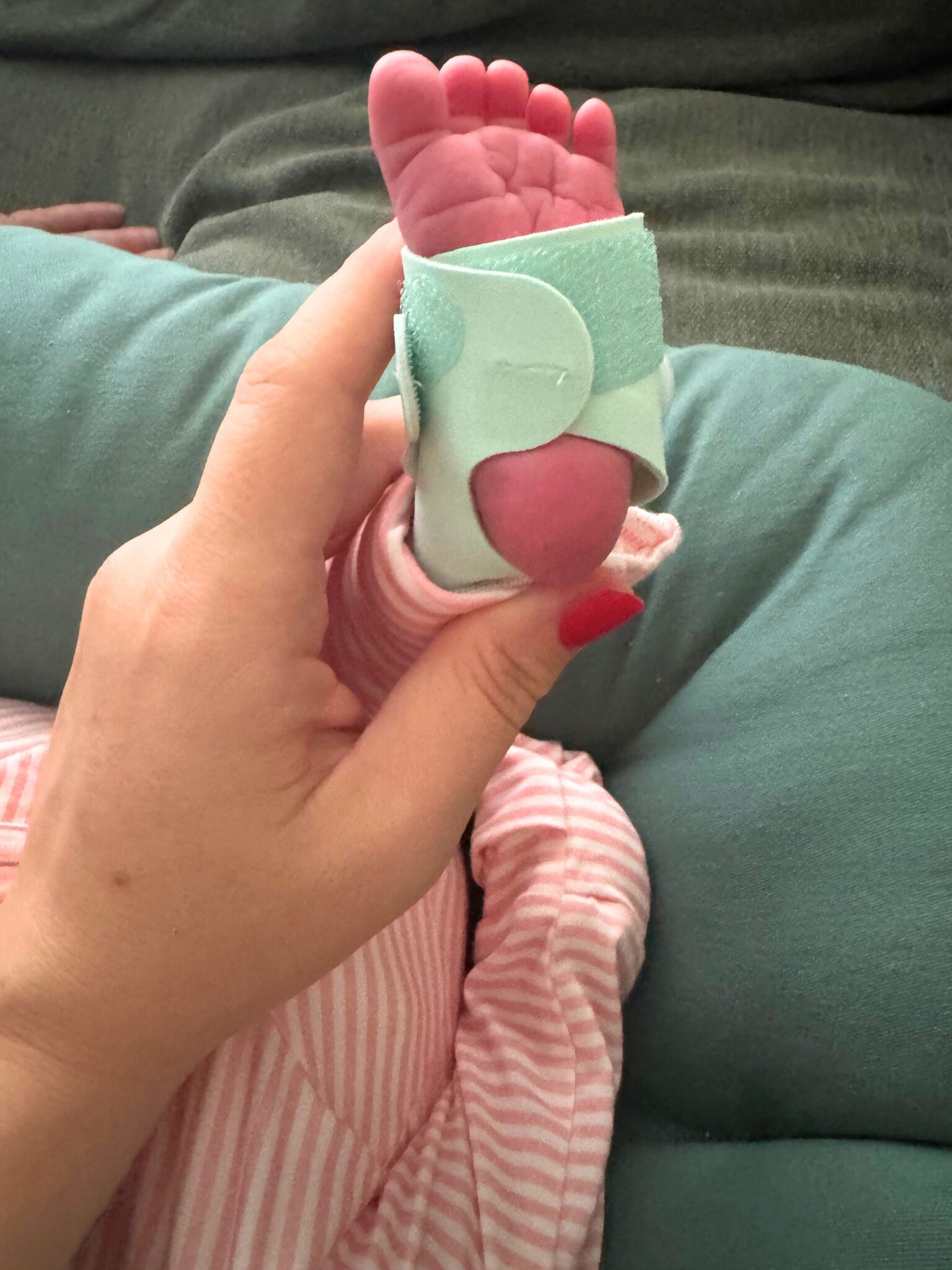 A hand holds a baby's foot with a mint green medical monitor wrapped around it; the baby is wearing pink striped pajamas.