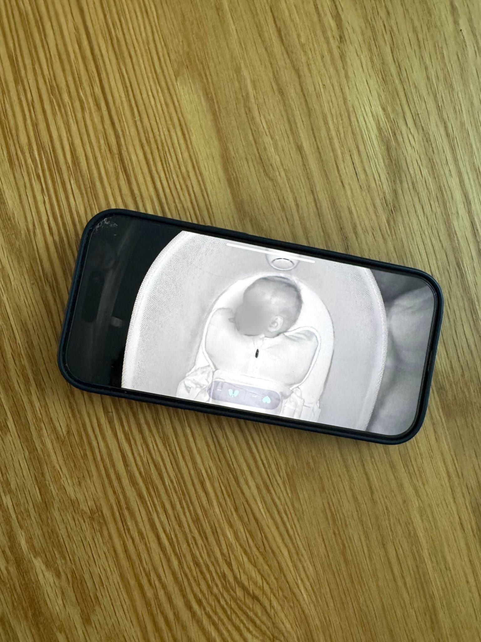 A smartphone on a wooden surface displays a grayscale video feed of a baby lying in a bassinet.