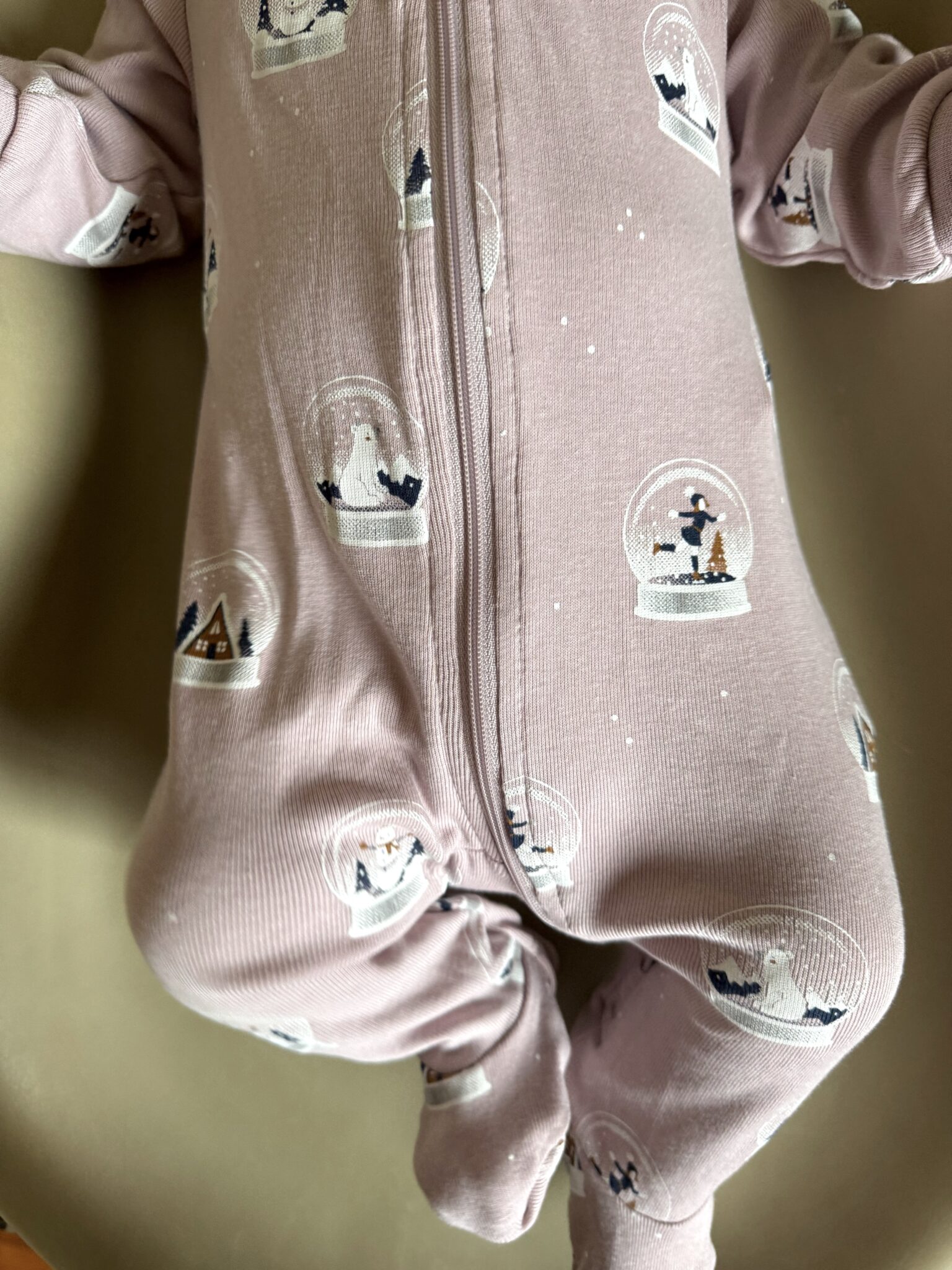 A baby wearing a light pink onesie with a zipper and winter-themed snow globe prints, shown from the neck down.