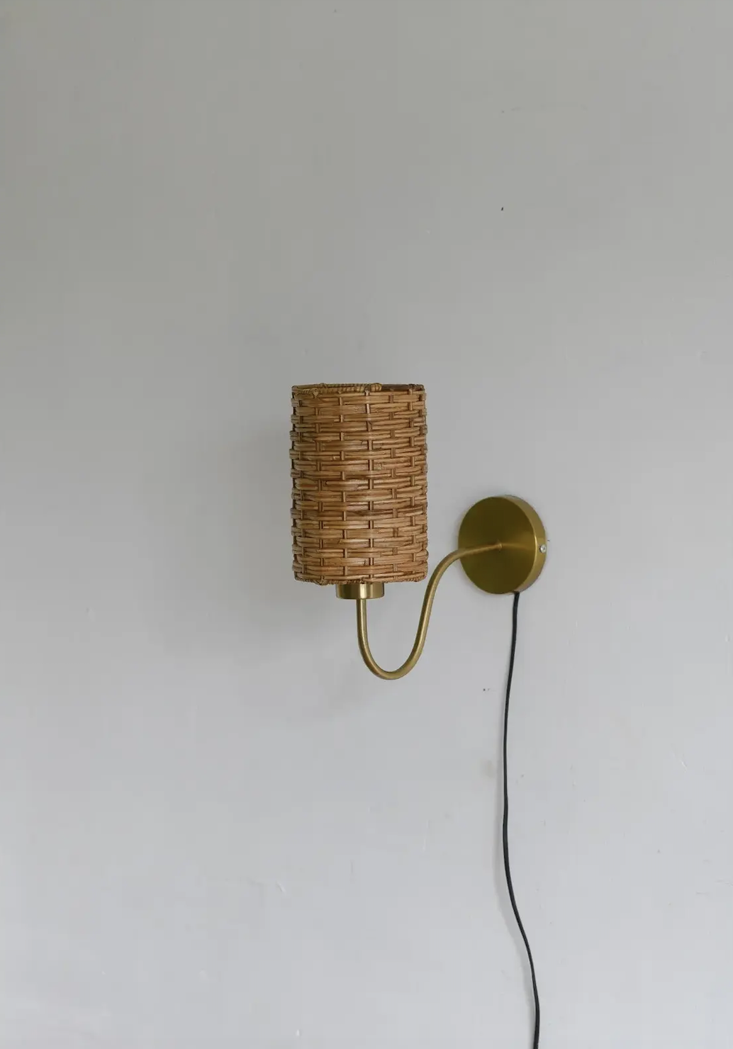 A brass wall-mounted lamp with a woven rattan shade and a visible black power cord against a plain light gray wall.
