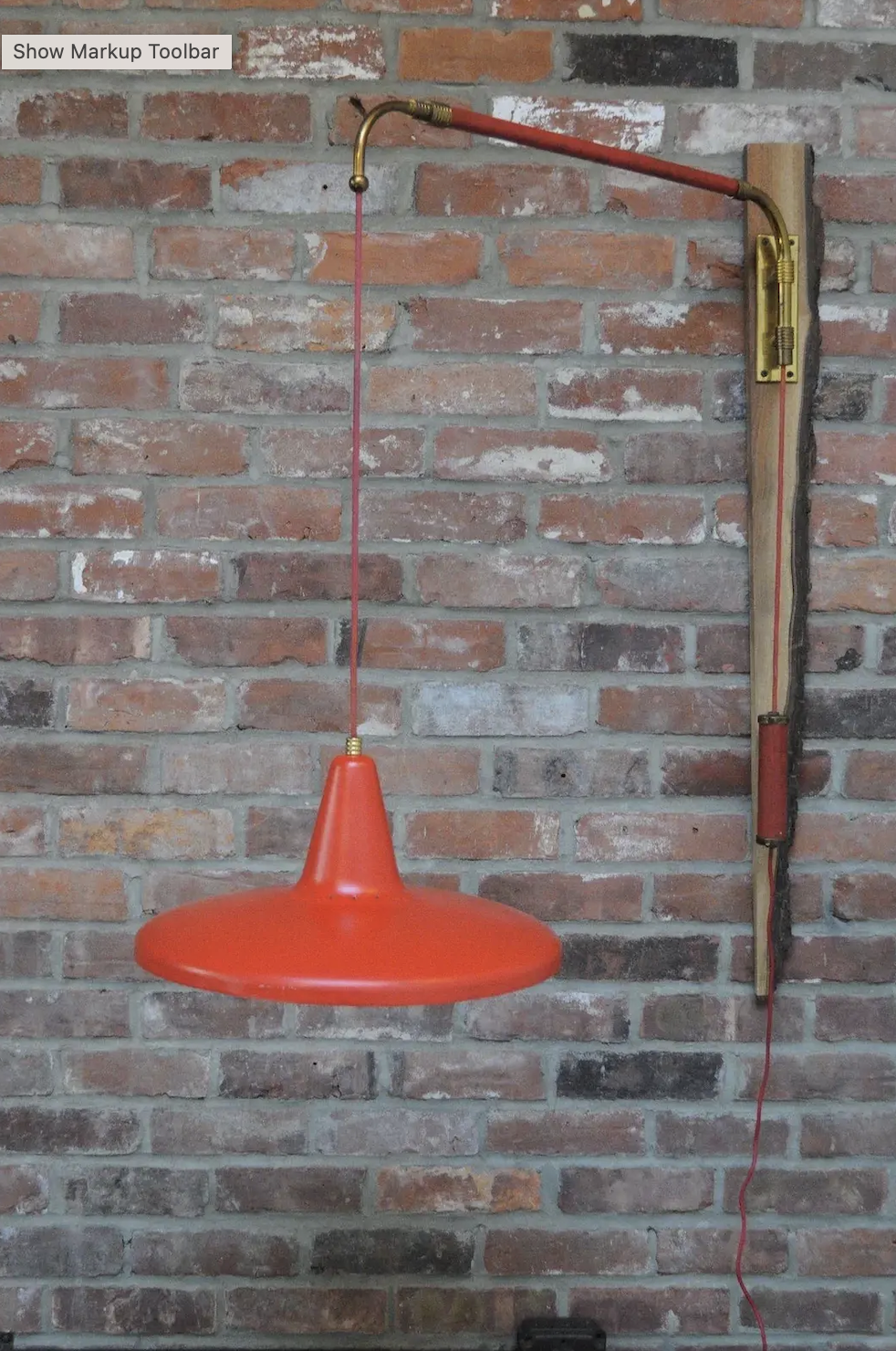A red pendant lamp hangs from a brass and wood wall-mounted arm against a red brick wall.