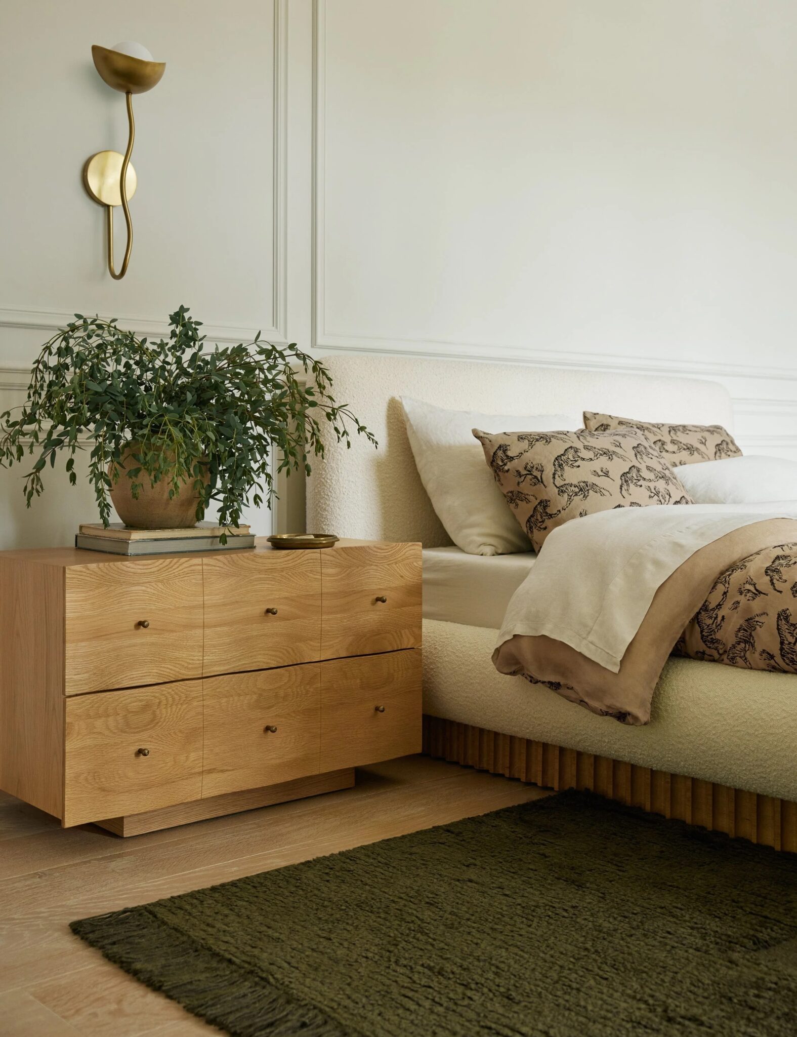 A modern bedroom with a wooden nightstand holding a potted plant, books, and a tray beside a bed with neutral bedding and patterned pillows.