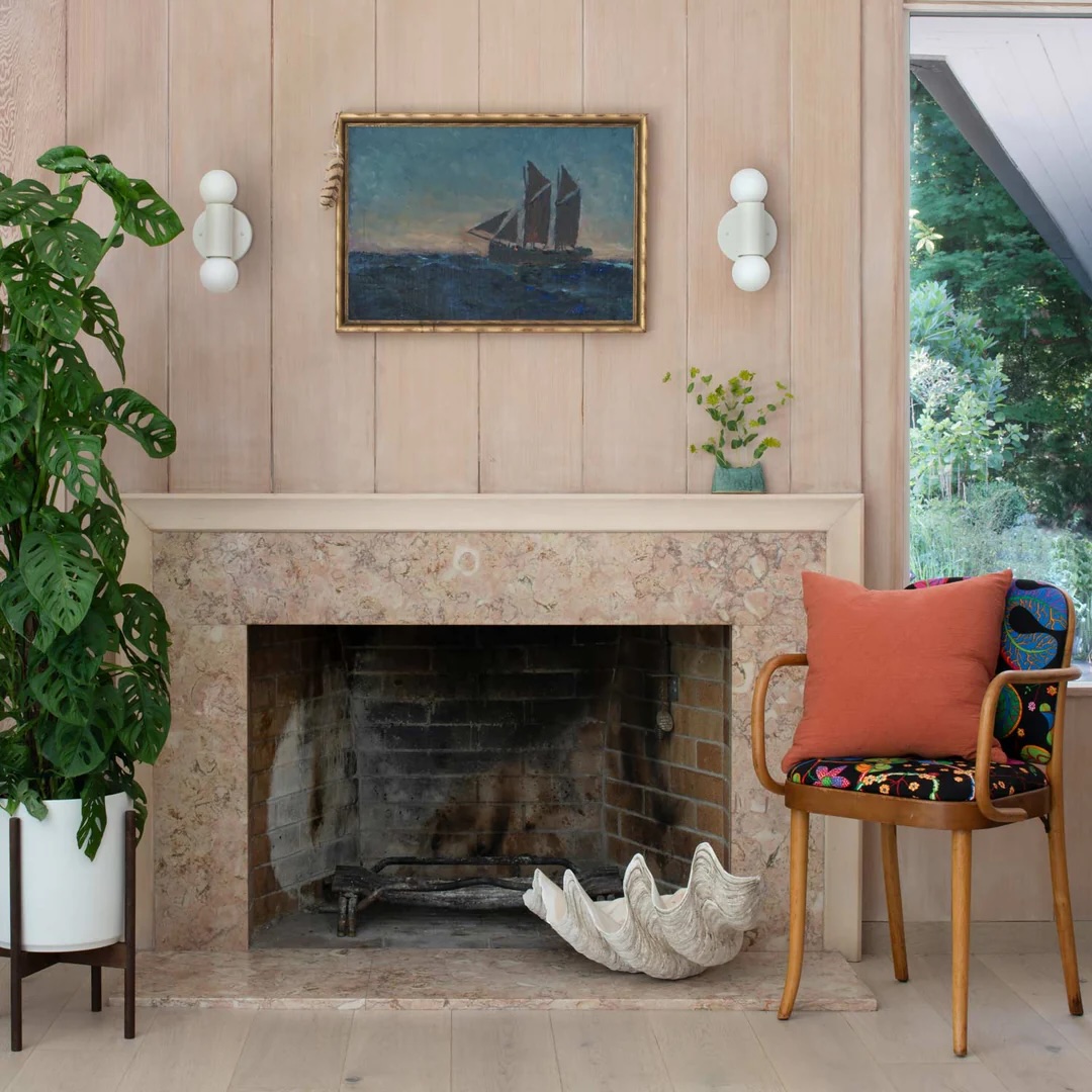 A marble fireplace with a large seashell in front, a potted monstera plant on the left, and a wooden chair with cushions on the right; a nautical painting hangs above the mantel.
