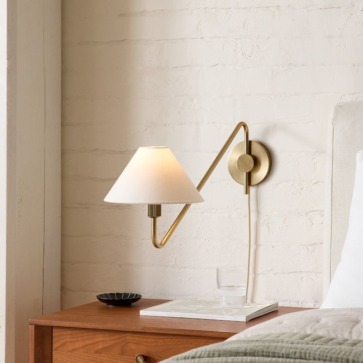 A brass wall-mounted lamp with a white shade is positioned above a wooden nightstand holding a small glass of water, a dish, and a marble tray, next to a bed with a light-colored headboard.