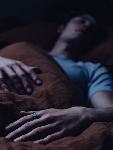 A person wearing a ring lies in bed under a brown blanket, with one hand resting on top of the blanket and the other partially visible.