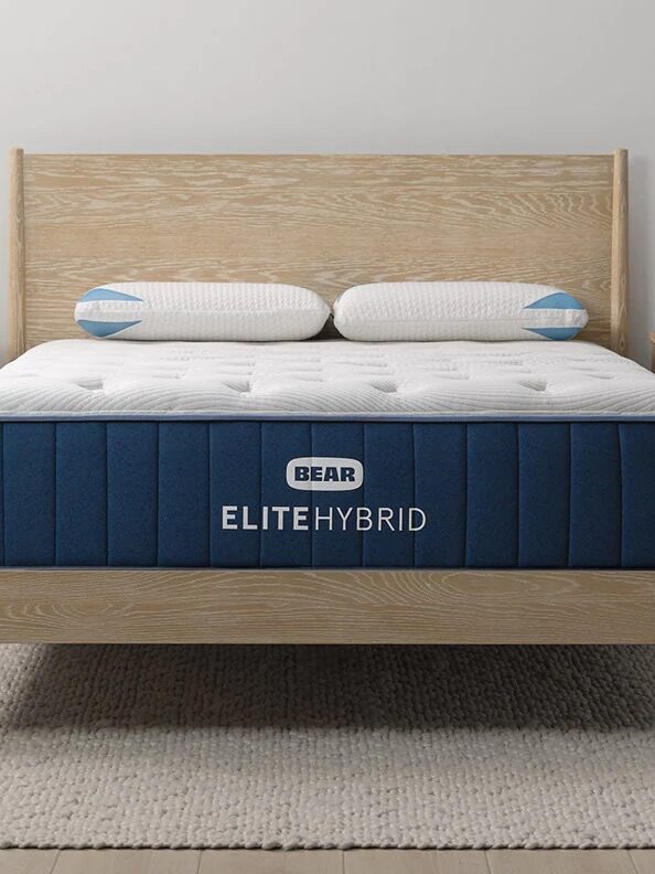 A neatly made bed with a blue and white Bear Elite Hybrid mattress, wooden headboard, and bedside tables; a sign reads “Upgrade to the Celliant Cover.”.