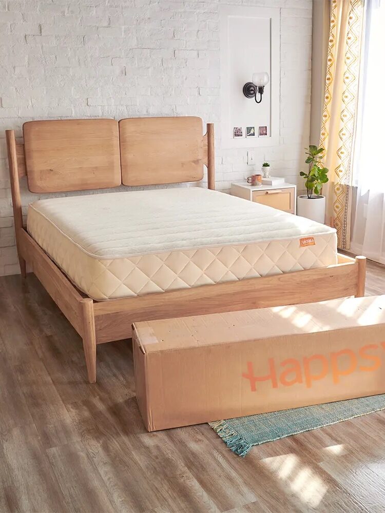 A wooden bed frame with a mattress is placed in a bright bedroom. A large cardboard box labeled "Happsy" sits at the foot of the bed on a blue rug.