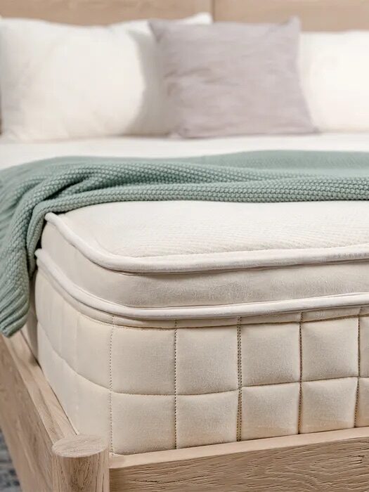 A close-up of a neatly made bed with a thick, beige mattress, white bedding, a light green blanket, and a wooden bed frame.