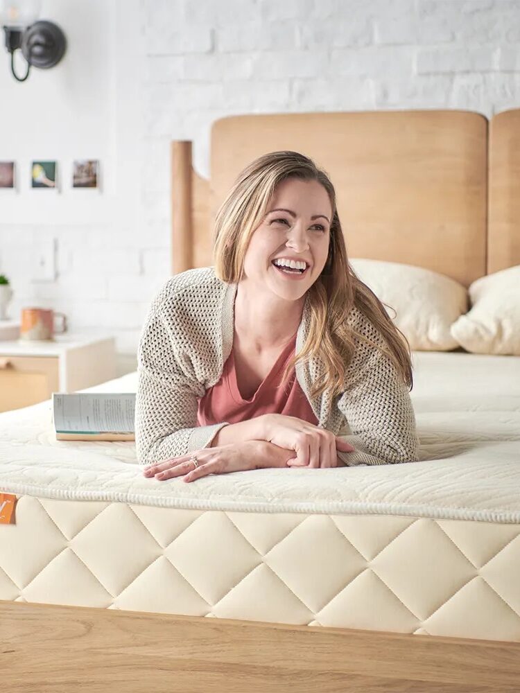 A woman lies on her stomach on a bed with a quilted mattress, smiling, with a book and a cup beside her in a bright bedroom.