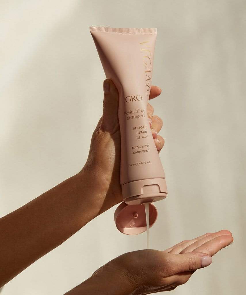 A hand dispenses clear shampoo from a pink VEGAMOUR GRO Revitalizing Shampoo bottle into the palm of another hand.