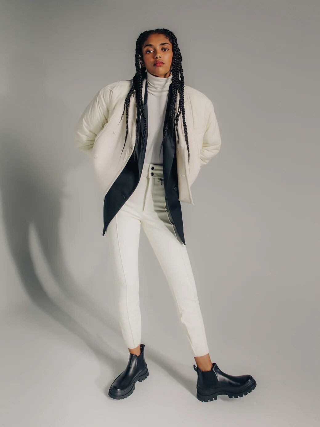 A person with braided hair poses in a light-colored jacket over a black blazer, white turtleneck, white pants, and black boots against a plain background.
