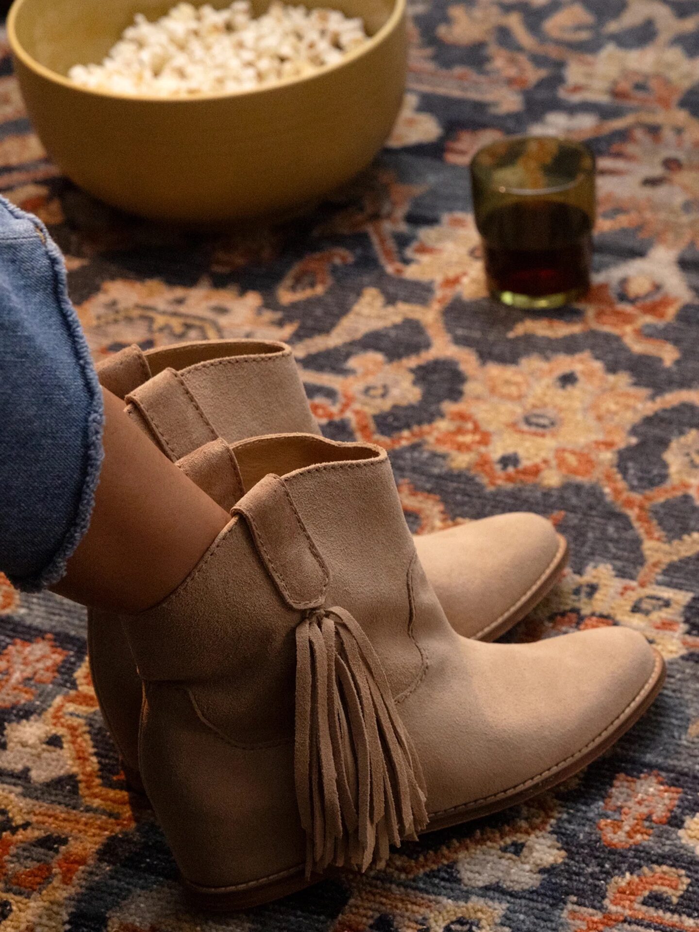 A person wearing tan suede boots with tassels sits on a patterned rug near a bowl of popcorn, a drink, and playing cards.