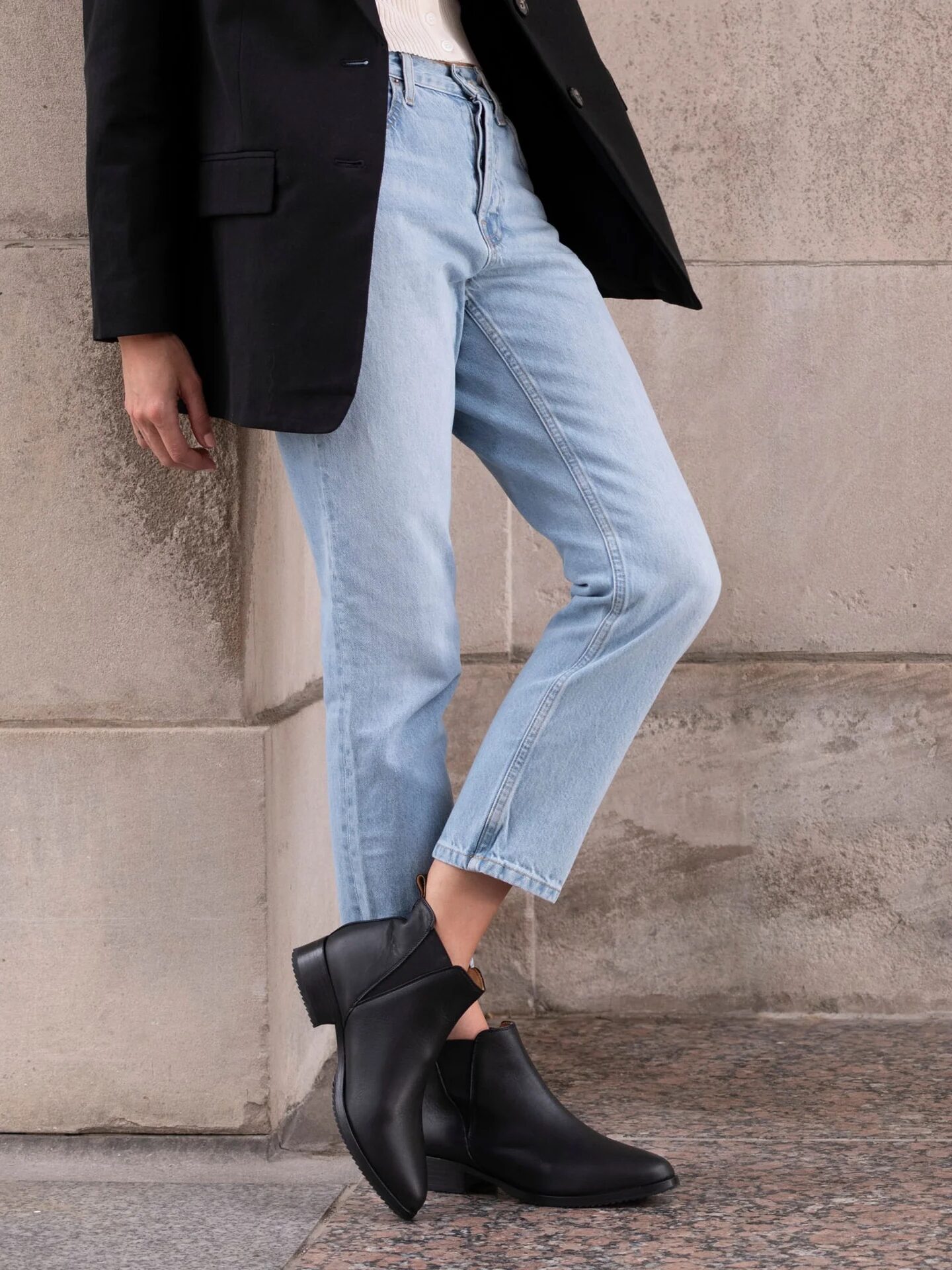 Person in a black blazer, light blue jeans, and black ankle boots stands against a stone wall with one leg crossed over the other.