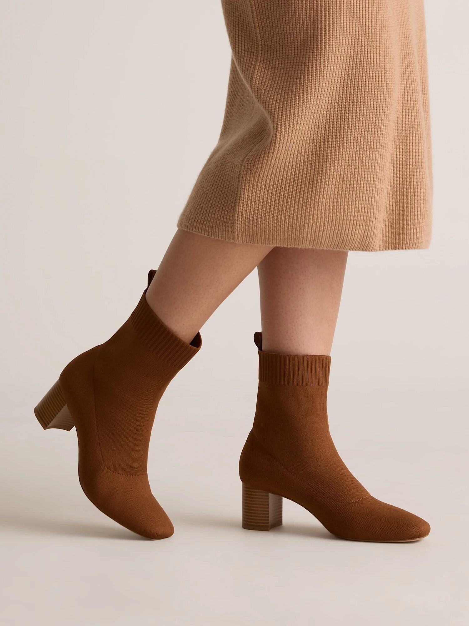 A person wearing a tan ribbed knit skirt and brown heeled ankle sock boots stands on a neutral background.