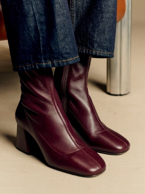 A person wearing dark jeans and burgundy heeled ankle boots sits on a chair with metallic legs.