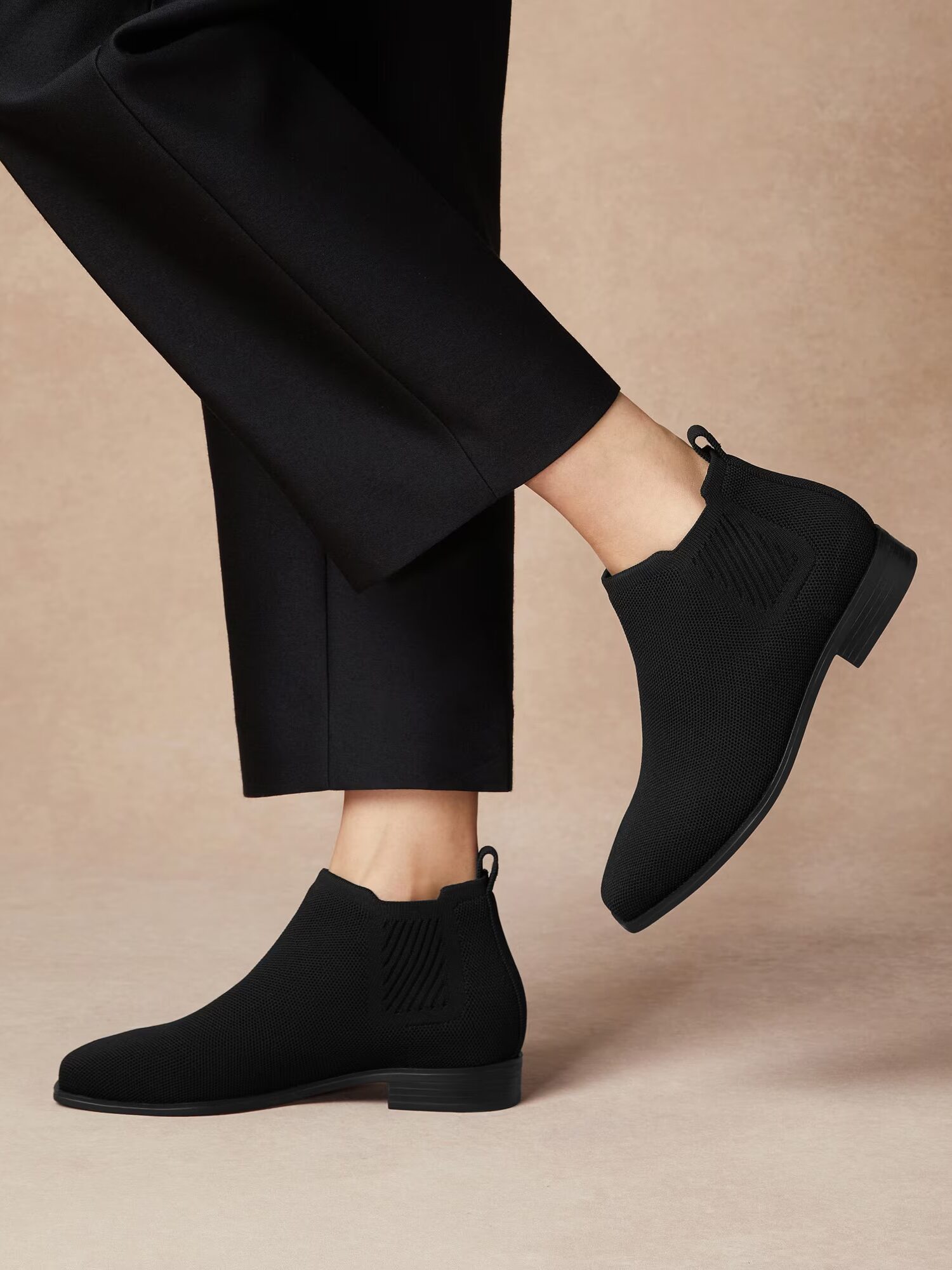 A person wearing black fitted pants and black ankle boots stands on a beige background, with one foot slightly raised.