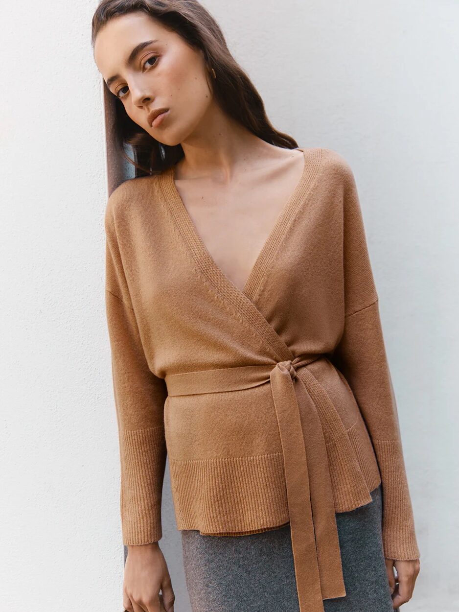 Woman wearing a tan wrap sweater with a tie waist and a gray skirt, standing against a white wall.