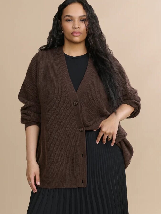A person wearing a brown button-up cardigan over a black top and pleated black skirt stands against a beige background.