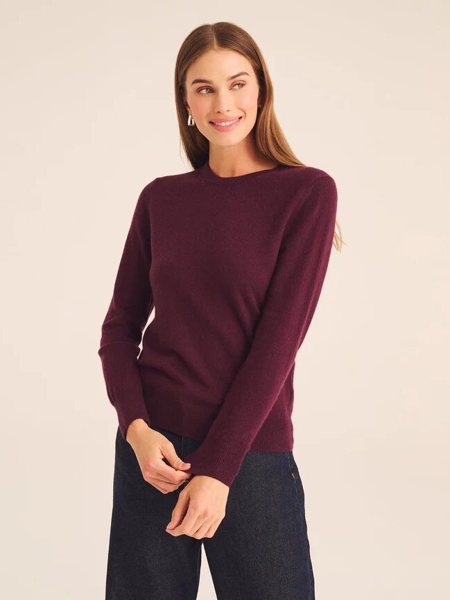 A woman with long brown hair wears a maroon sweater and dark pants, standing against a plain light background.