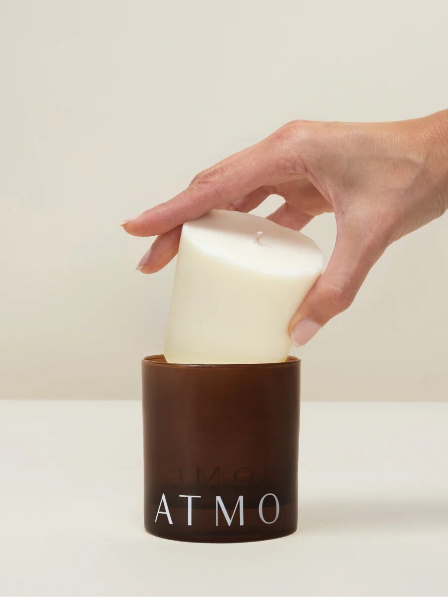 A hand places a white pillar candle into a brown glass holder labeled "ATMO" against a plain background.