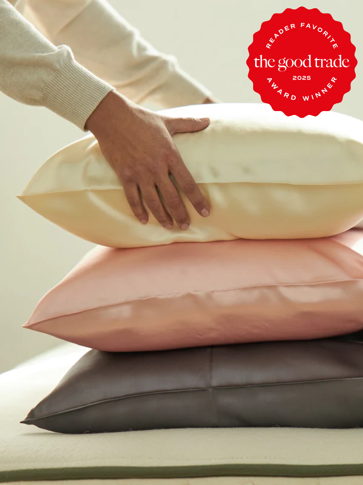 A person places a hand on a stack of three satin pillows in cream, pink, and dark gray. A red badge in the top right corner reads “the good trade 2025 Award Winner.”.