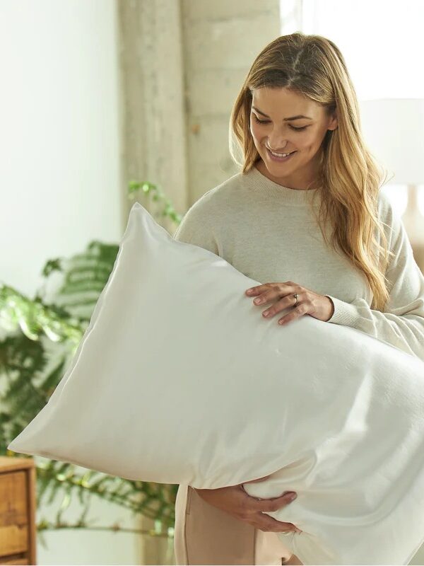 A woman stands indoors, holding a large white pillow and smiling. There are plants and a lamp in the softly lit background.