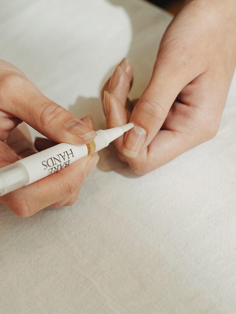 A person applies a product labeled “Bare Hands” to their fingernail using a pen-like applicator, with hands resting on a white surface.