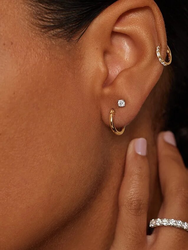 A close-up of a woman's ear with three earrings—two gold hoops and a small diamond stud—and a hand wearing a gold and diamond ring.