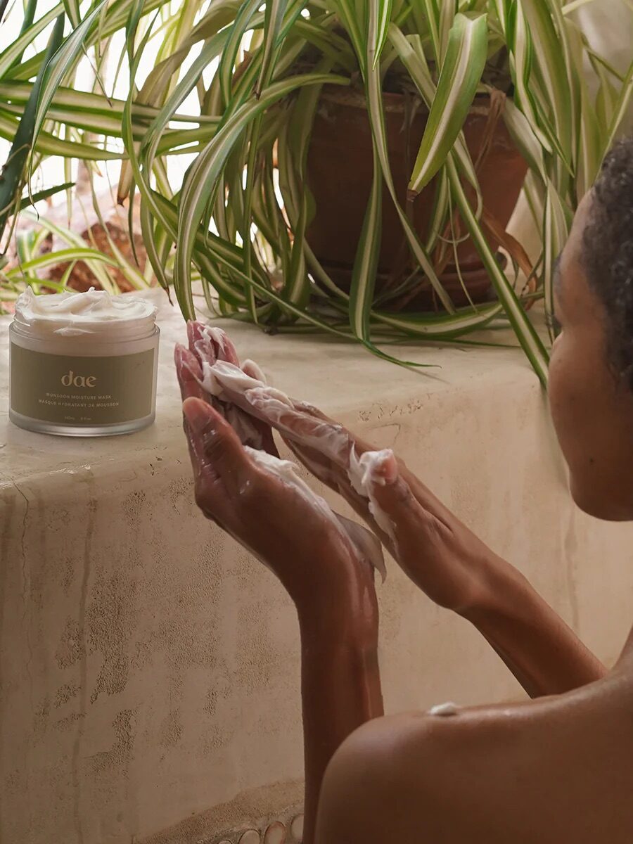 A person applies a creamy product from a jar labeled "dae" onto their arms while sitting near potted plants on a ledge.