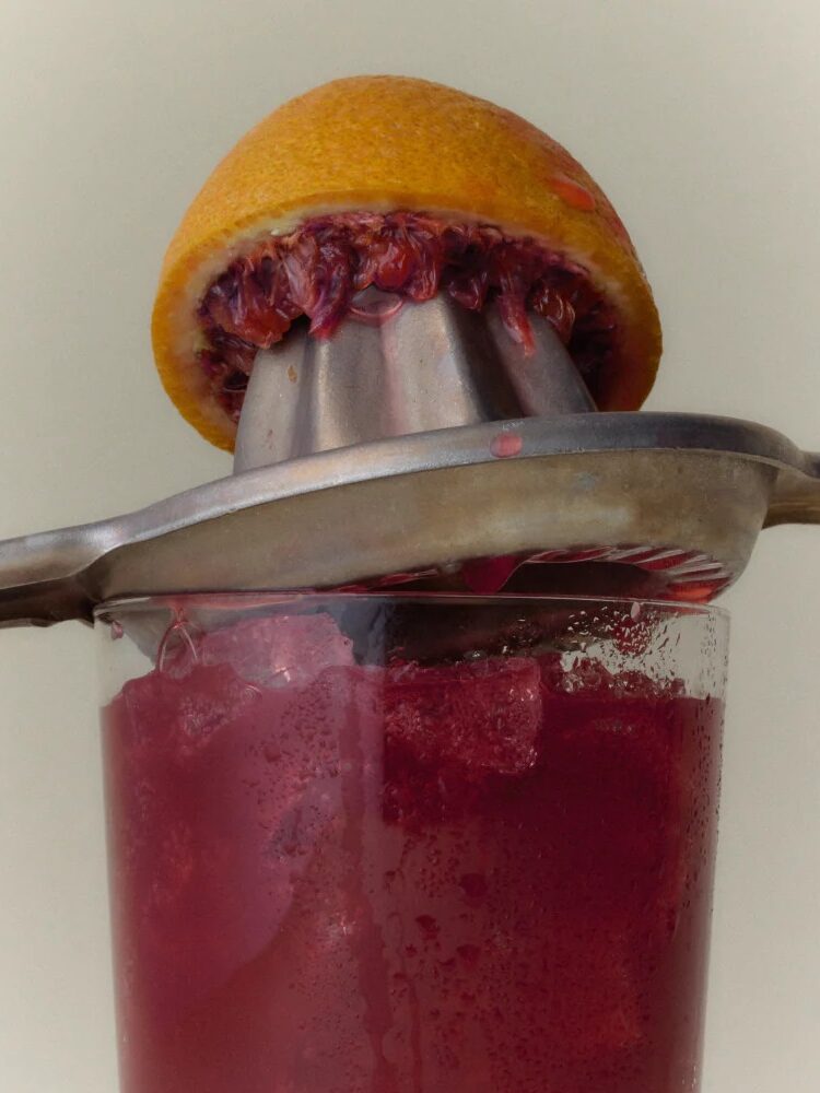 A metal citrus juicer presses a halved blood orange over a glass filled with dark red juice and ice.