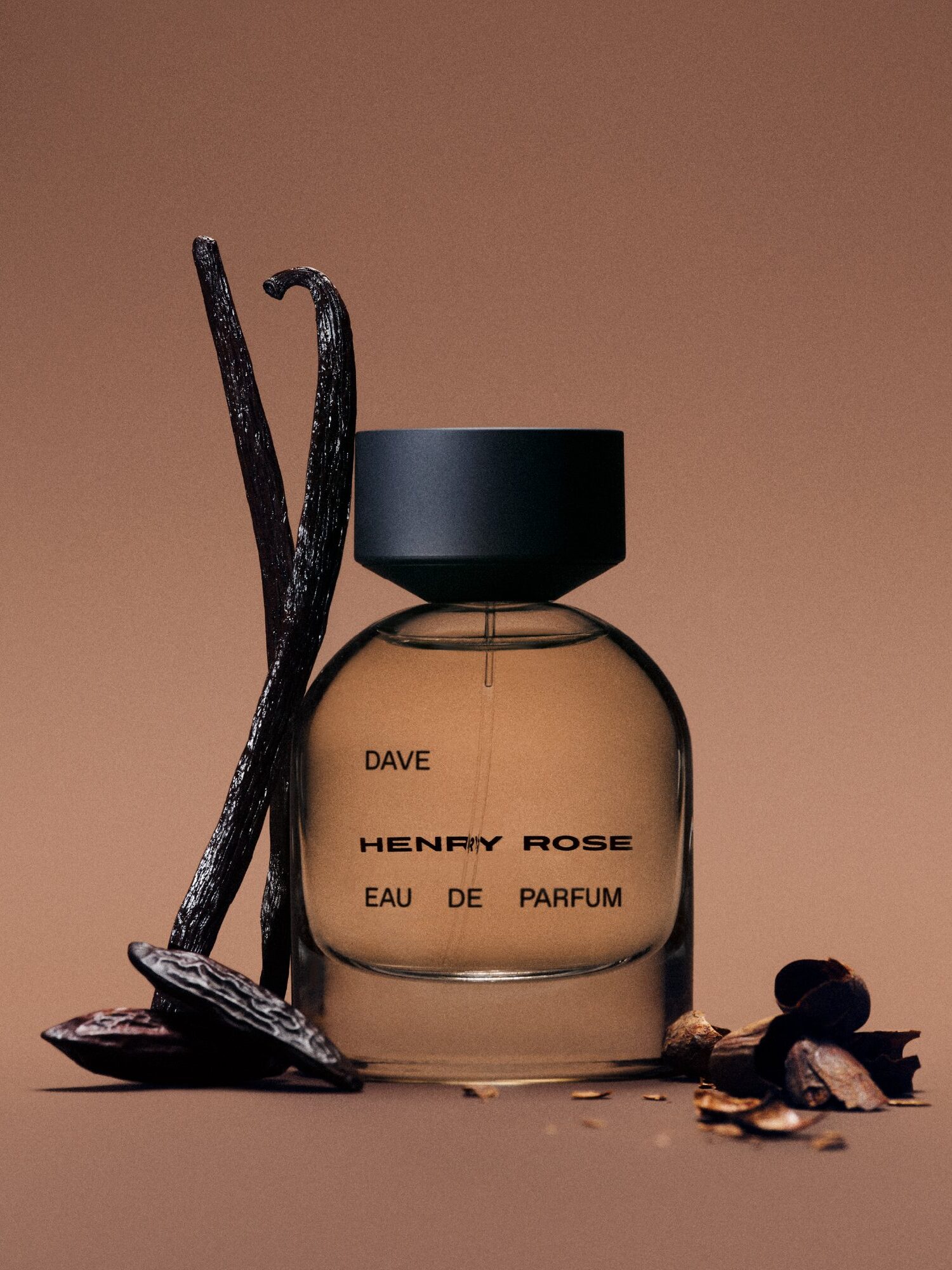 A bottle of Henry Rose "DAVE" Eau de Parfum stands upright, flanked by vanilla beans and scattered dried plant material on a brown background.