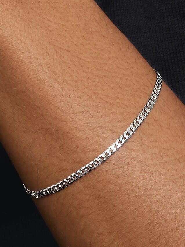 A close-up of a person's wrist wearing a thin silver chain bracelet against a dark background.