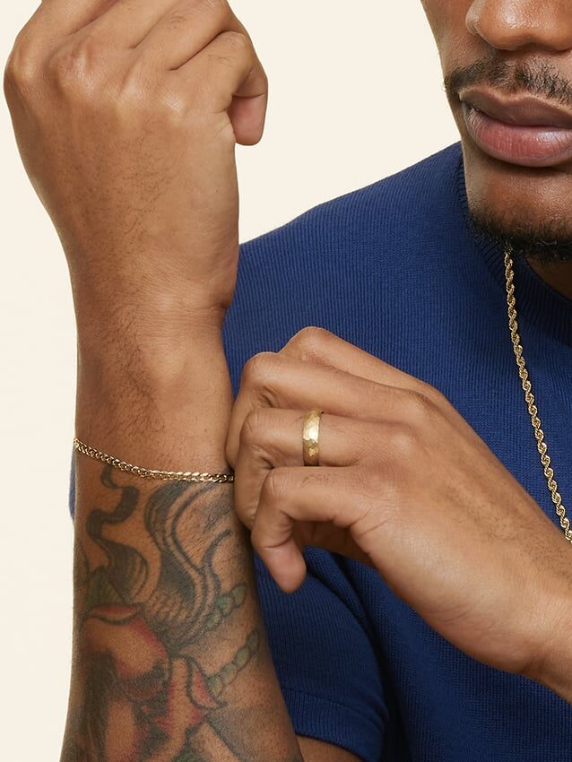 A man in a blue shirt displays a gold chain bracelet and ring on his tattooed wrist and hand.