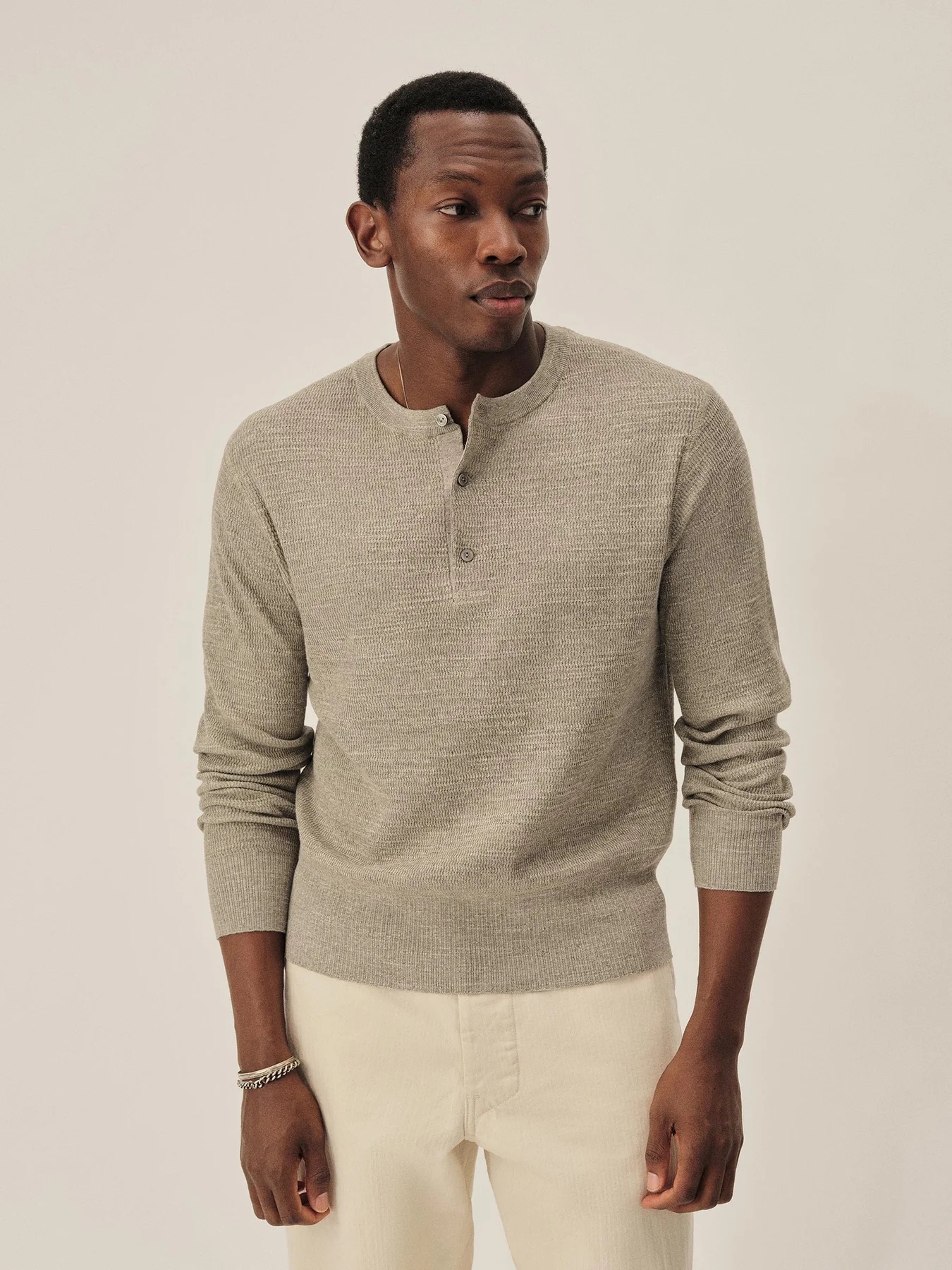 A man wearing a light gray henley sweater and beige pants stands against a plain, light background.