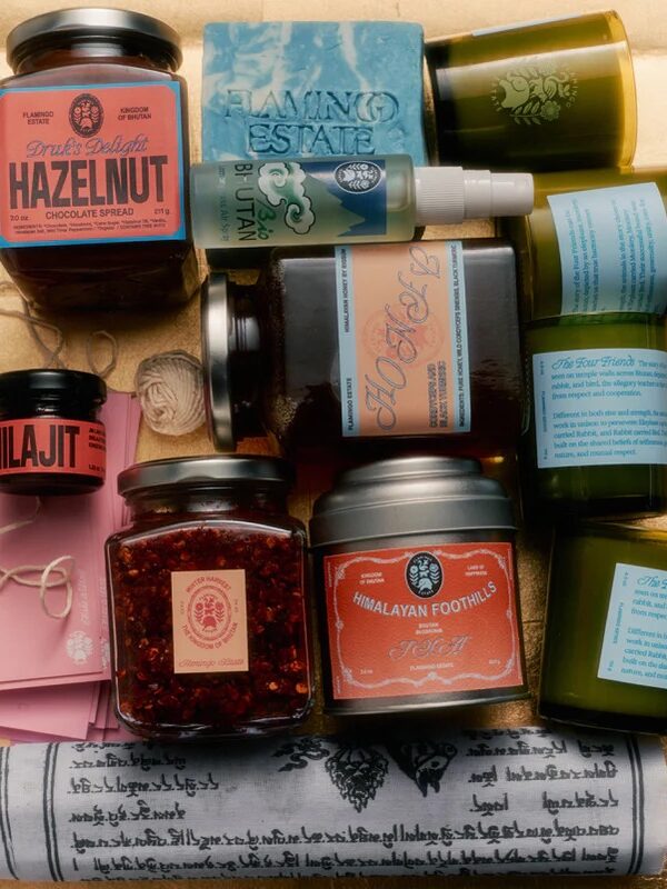 Assorted jars, candles, soaps, and bottles are arranged on a gold surface along with printed paper cards and a rolled fabric item featuring printed text.