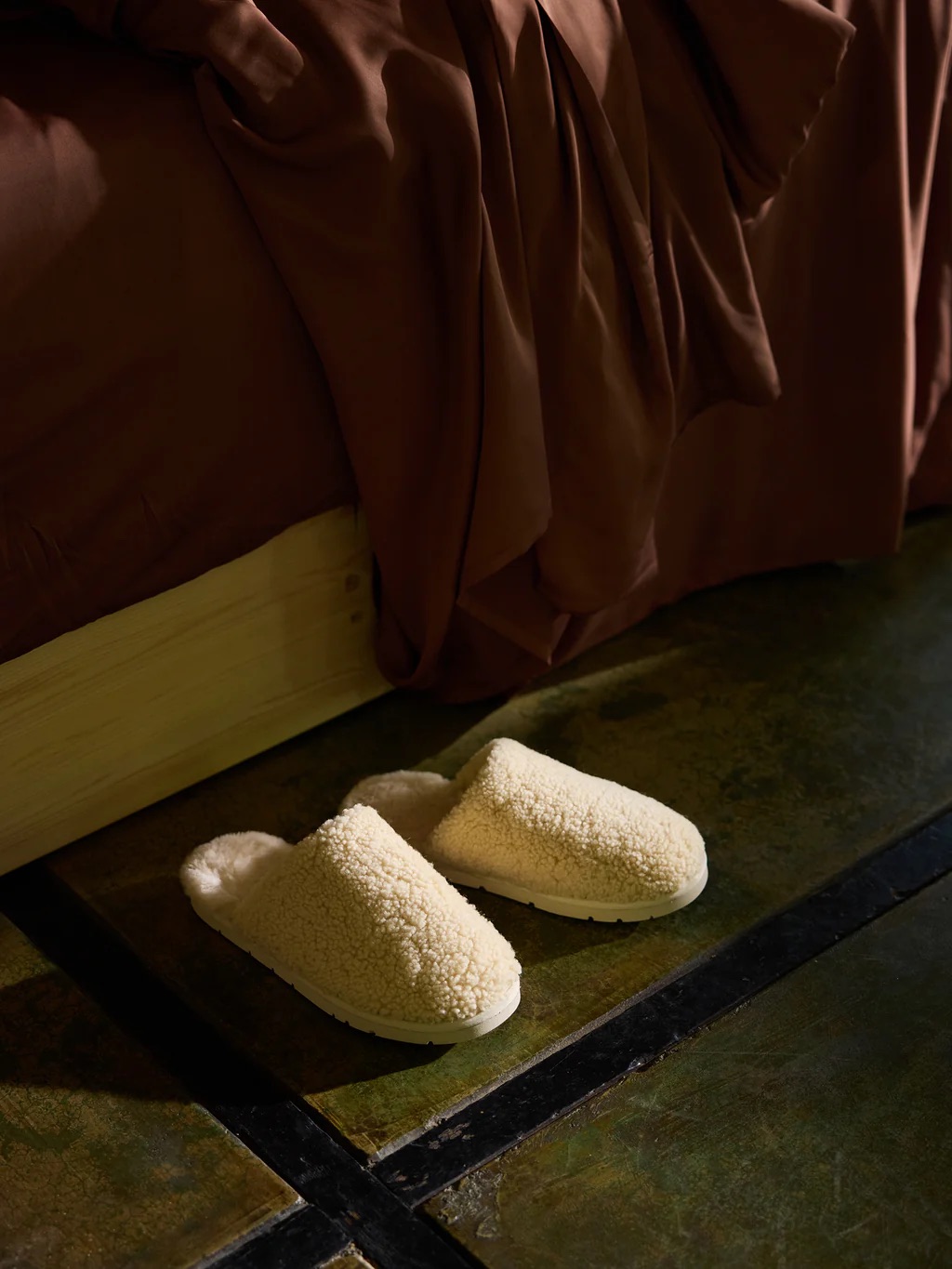 A cozy pair of cream-colored slippers rests on the polished floor, nestled beside a bed draped in soft brown bedding.