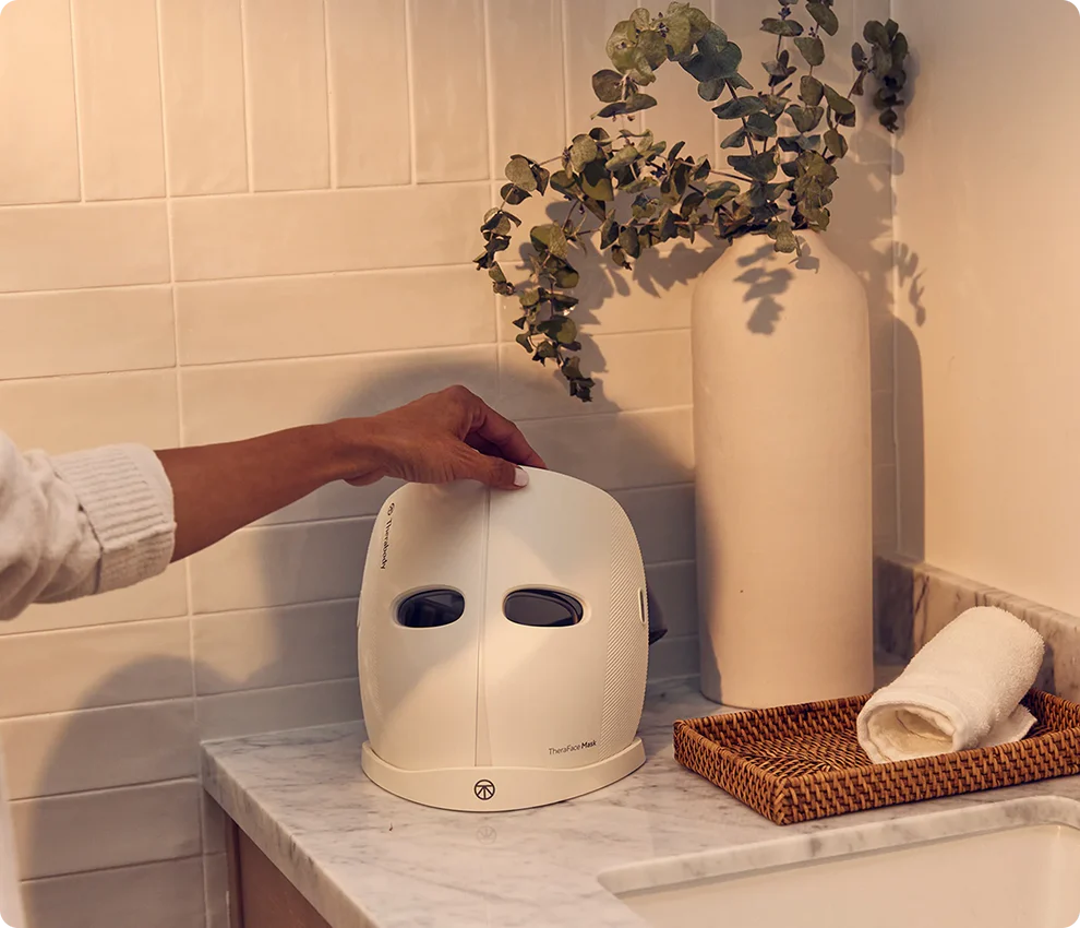 A person places a white LED face mask on a marble bathroom counter next to a vase with green foliage and a woven tray holding a rolled towel.
