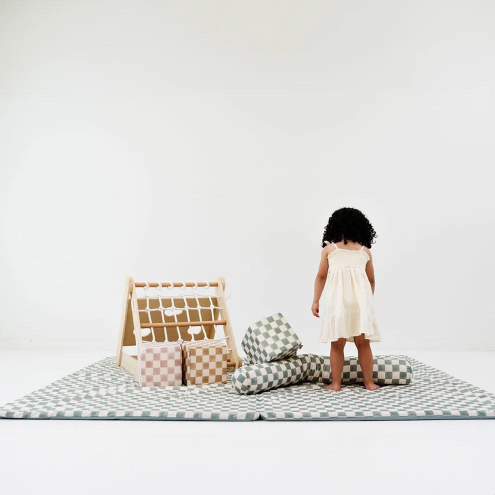 A young child in a white dress stands on a checkered playmat with soft blocks and a wooden activity toy in a minimal, white room.