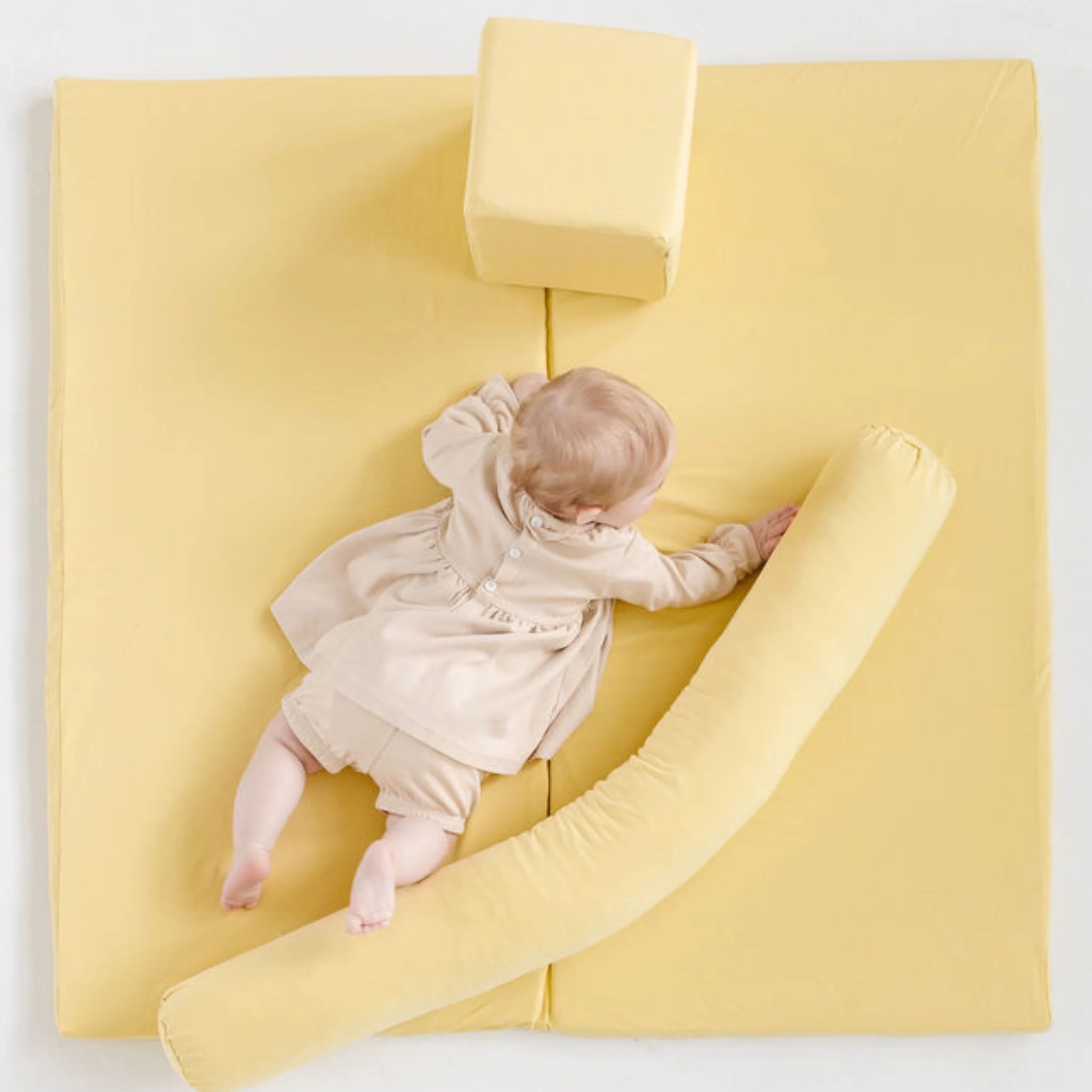 A baby in light-colored clothes lies on a yellow padded play mat with a cylindrical and a cube-shaped cushion.
