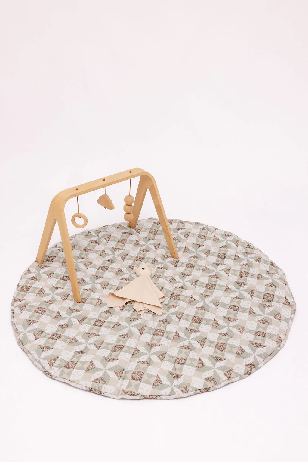 A wooden baby gym with hanging toys stands on a circular patterned play mat, with a beige cloth doll placed nearby.